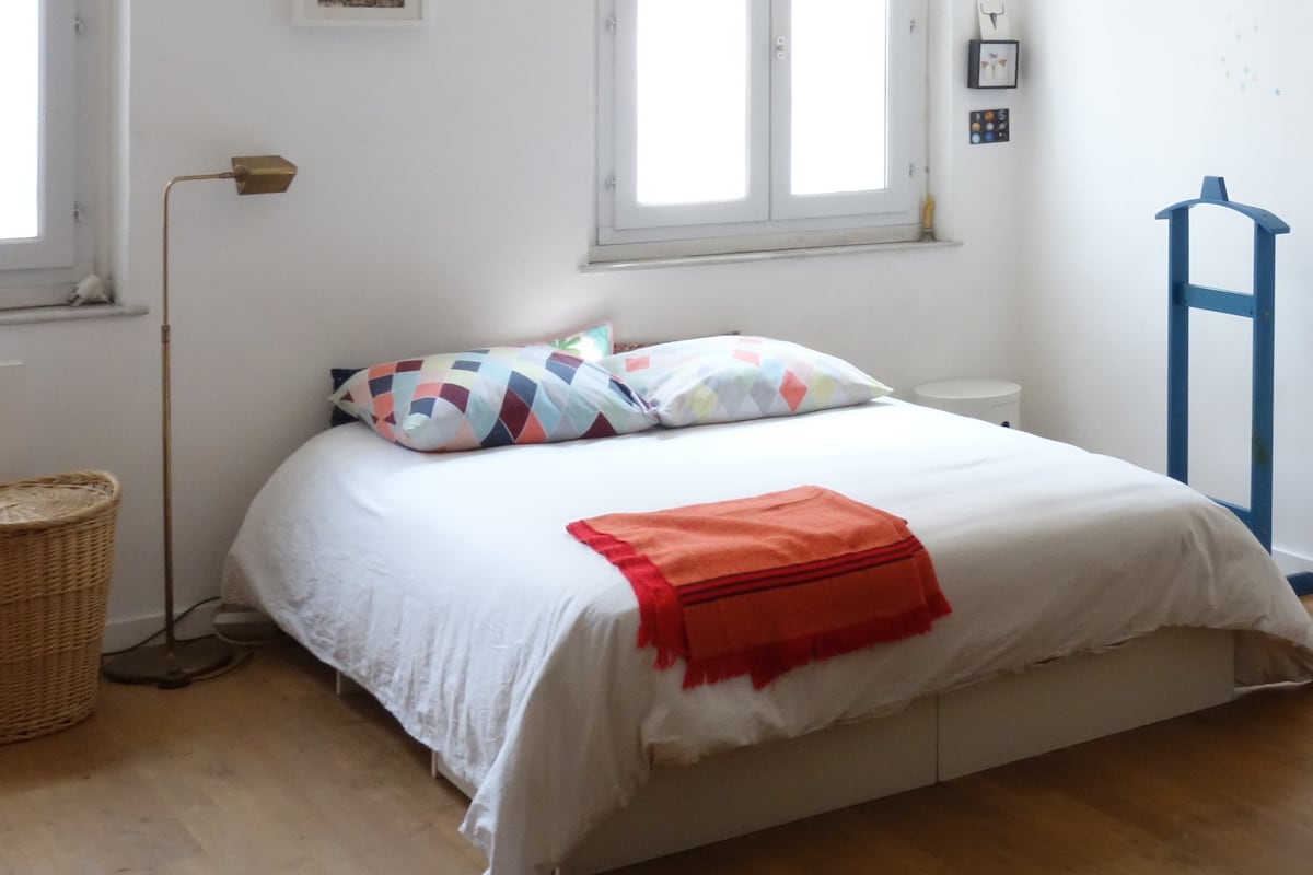 A spacious bed is centered in a bright room, adorned with colorful patterned pillows. Natural light enters through several windows, illuminating the warm wood flooring. A stylish lamp and a woven basket are positioned nearby, adding to the functional decor.