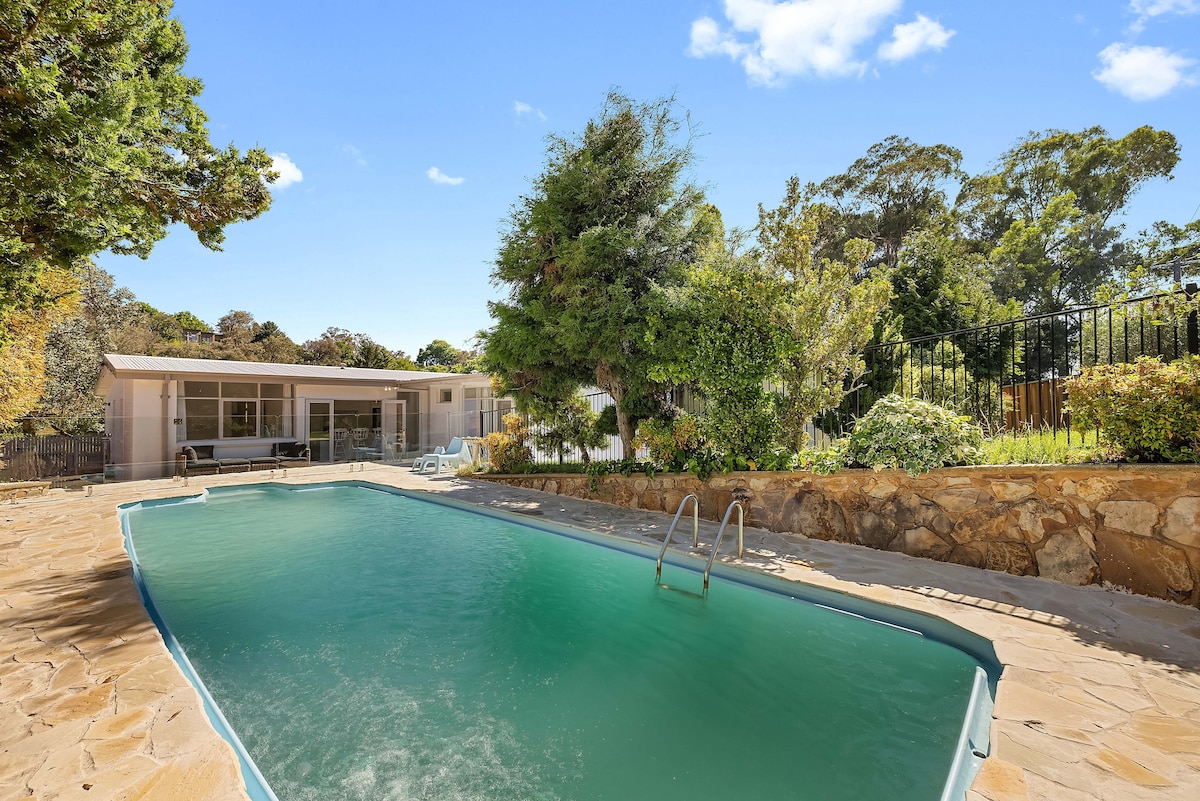 The image captures a private pool surrounded by a stone patio, framed by lush greenery. The pool's water glistens in natural light, reflecting the clear blue sky. In the background, a modern home is partially visible behind a hedge, creating a serene outdoor retreat.