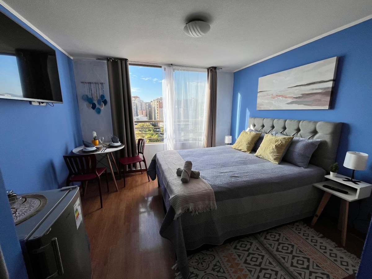 A bright and inviting home studio features a spacious bed with decorative pillows, accompanied by a small dining table for two. A large window offers a view of the city, while blue walls add a calm ambiance. A small fridge is positioned in the corner.