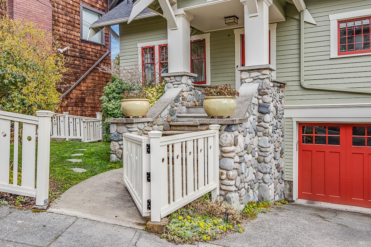 The exterior entrance of the 1914 bungalow is showcased, featuring decorative stonework and a charming railing. Two large, rounded planters rest on the stone steps, which lead to a warm-colored door. The pathway is bordered by manicured greenery, enhancing the inviting atmosphere.