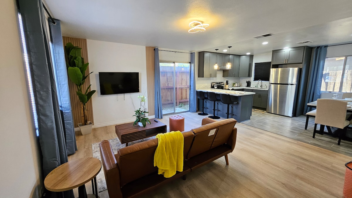 A spacious living area is highlighted by a comfortable brown leather sofa and a wooden coffee table. A kitchen with modern appliances is visible in the background, complemented by a dining area featuring a round table. Natural light filters through large windows covered by light-colored curtains.