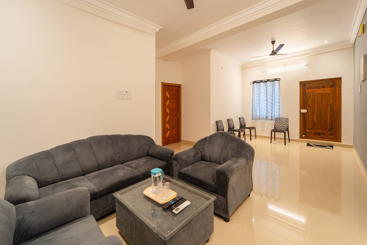 A living area features a plush gray sectional sofa and matching armchair arranged around a glass coffee table. Four dining chairs are positioned in the background. Natural light filters through a window adorned with light blue curtains, enhancing the spacious feel of the room.