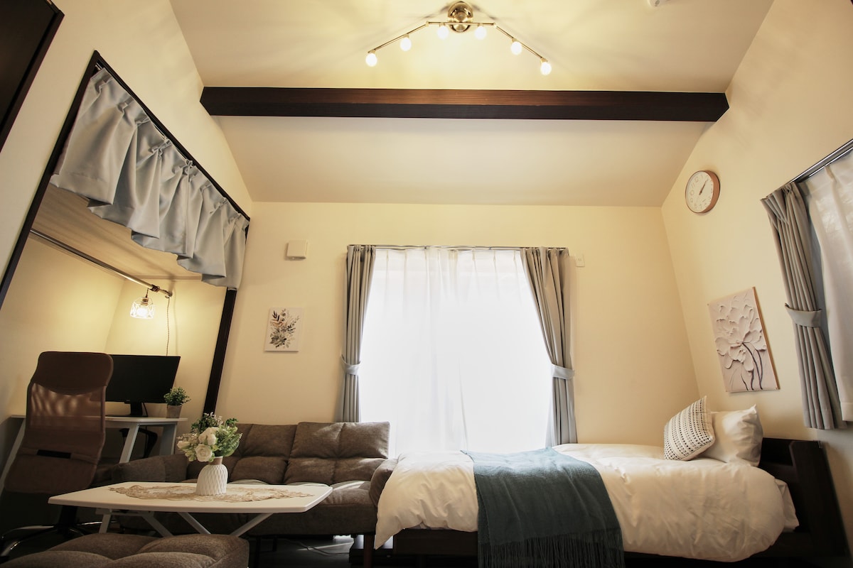 A compact living space features soft natural light streaming through two large windows adorned with light curtains. A comfortable sofa is positioned near a small coffee table, while a spacious bed is dressed in fresh linens. A work area with a desk is visible in the background.