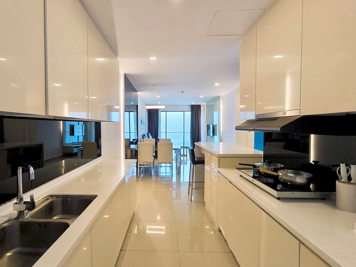 A modern kitchen space features sleek white cabinetry and a minimalist design. A functional countertop is complemented by a double sink and a stove area. Natural light filters through a window, illuminating the open layout that connects to a dining space visible in the background.