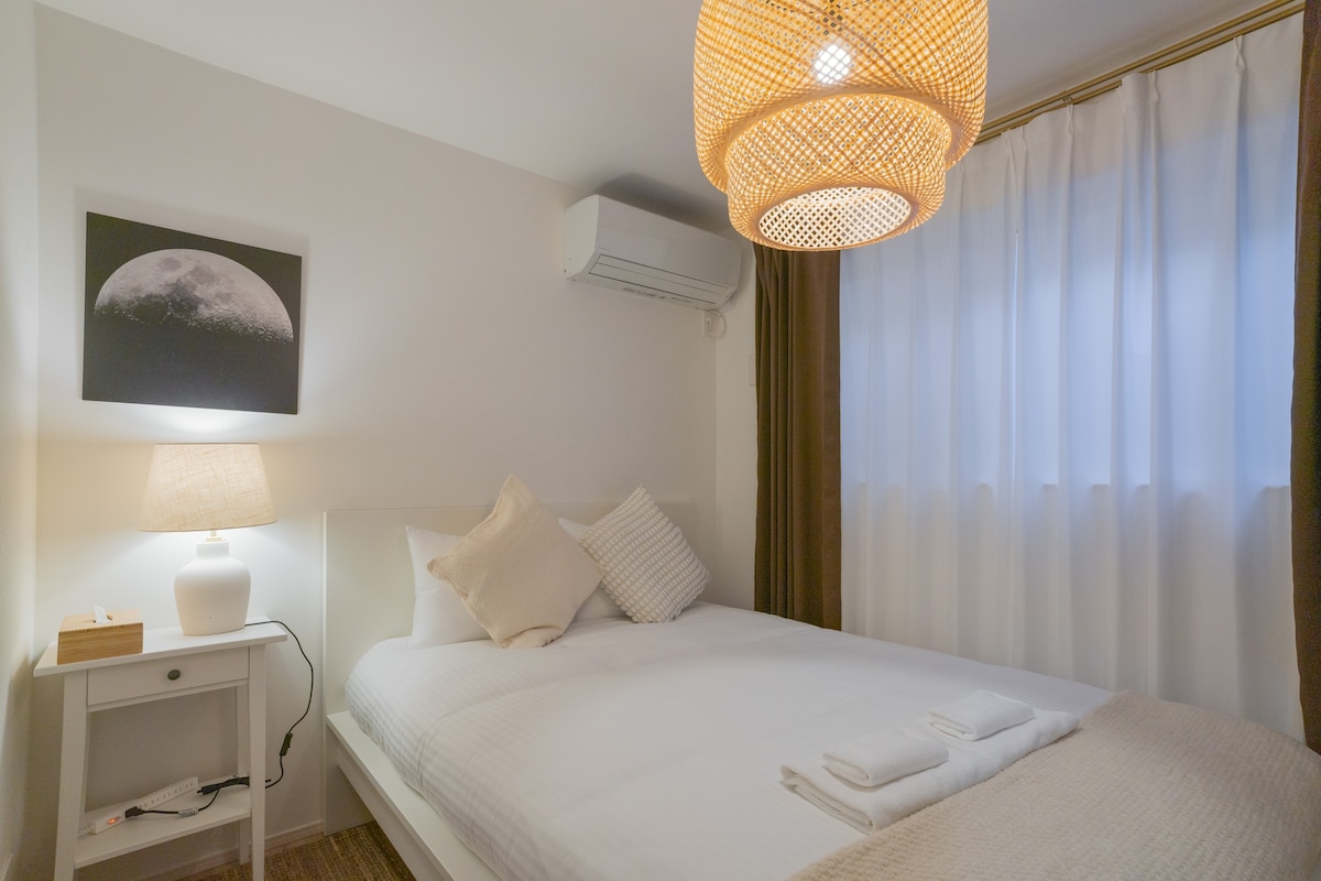 A well-lit bedroom features a spacious bed dressed in crisp white linens, complemented by a soft throw. A round pendant light hangs overhead, and a bedside lamp illuminates the area. Natural light filters through sheer curtains, enhancing the cozy atmosphere.