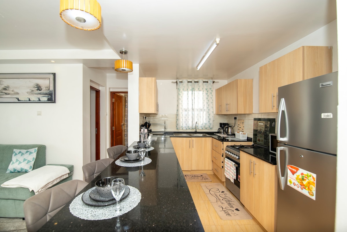 A modern kitchen is shown with light wooden cabinets and dark granite countertops. Four stylish bar stools are positioned at a counter, while various kitchen appliances and utensils are neatly arranged. Natural light fills the room through a window adorned with light curtains.