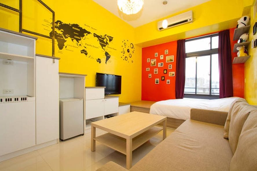 A bright and colorful room features a yellow wall with a world map and a red accent wall adorned with framed photos. A spacious bed is positioned next to a large window with dark curtains. A neutral-toned sofa and a small coffee table are arranged in the living area.