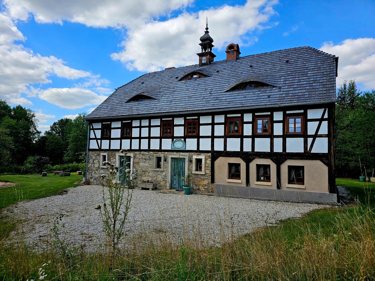 A charming two-story structure is presented with a mix of wooden beams and light-colored walls. The sloped roof features vented gables. A spacious grassy area is visible in front, complemented by a gravel pathway leading to the entrance, surrounded by natural greenery.