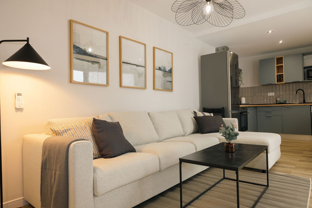 A modern living area is presented with a spacious light-colored sectional sofa, accompanied by decorative cushions. Above, three framed artworks adorn the wall. A sleek black coffee table sits in front of the sofa, complemented by a small potted plant. A stylish light fixture adds illumination.