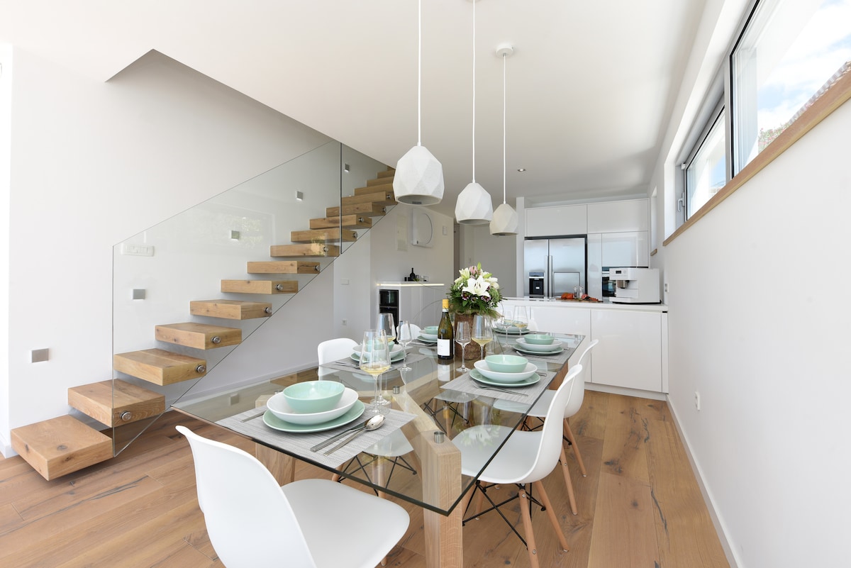 A modern dining area features a glass table set for six, surrounded by white chairs. Light wood floors complement the sleek design. Natural light streams through large windows, illuminating the space. A stylish staircase with a glass railing leads to the upper level, enhancing the openness.