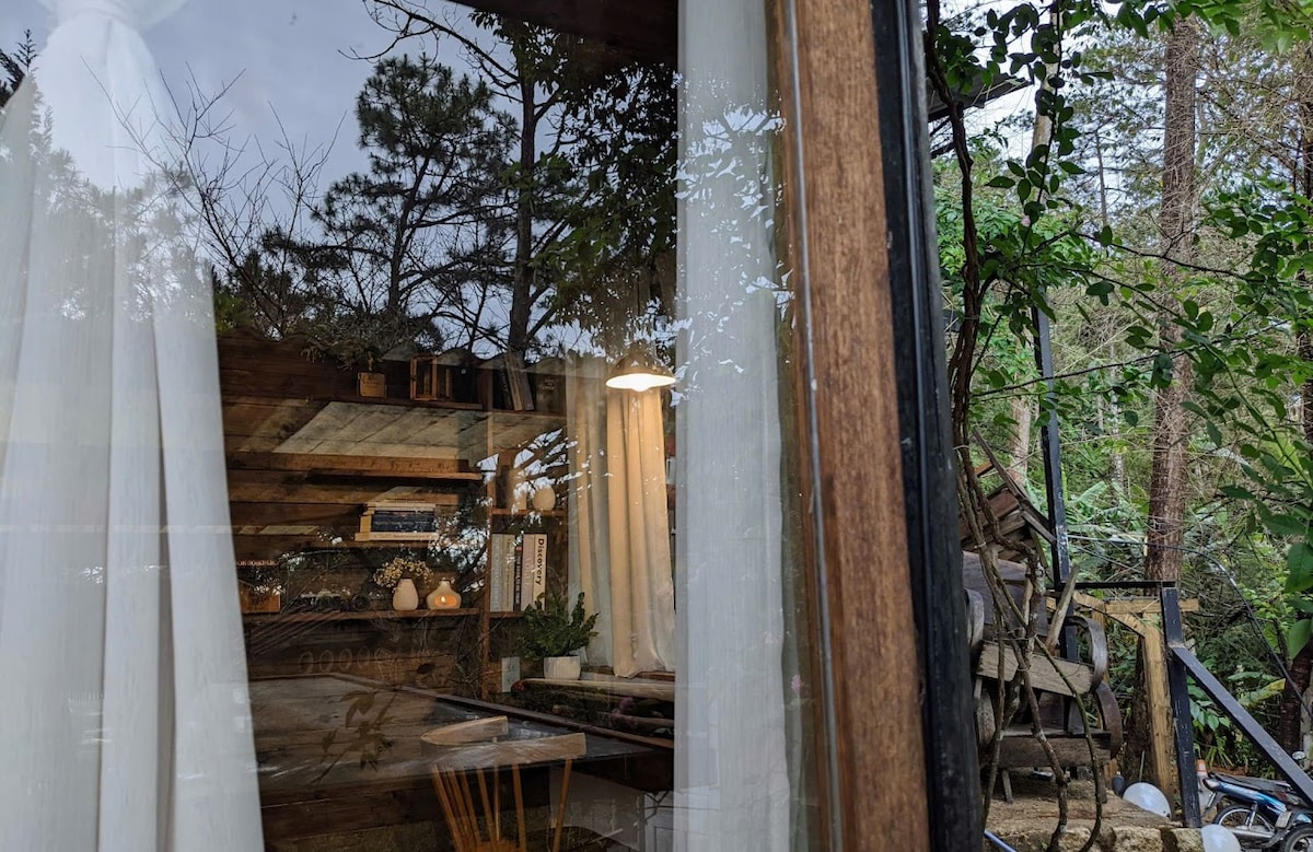 A cozy interior is visible through a large glass window, showcasing a wooden table and minimalist decor. Soft white curtains frame the glass, allowing natural light to enhance the warm tones of the wood. Lush greenery outside is partially seen, creating a connection with nature.