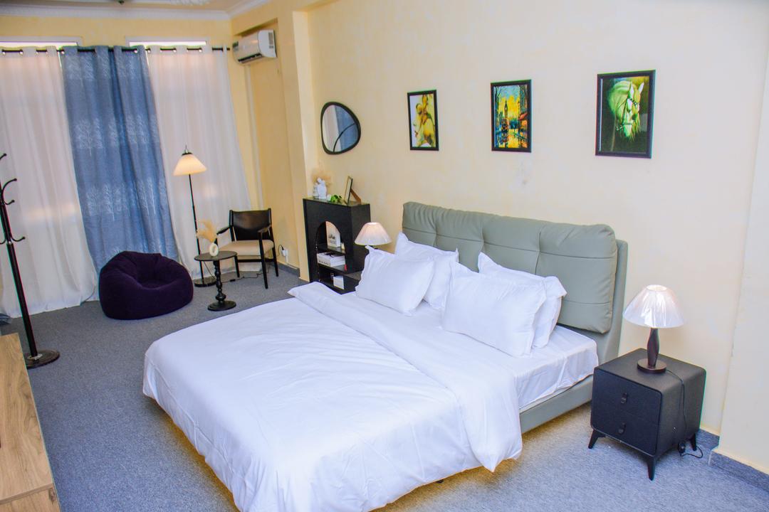 A calming bedroom features a spacious bed with crisp white linens. Two framed artworks are displayed on the wall above a dark dresser. A cozy armchair and a bean bag add seating options, while soft light from a bedside lamp creates a serene atmosphere.