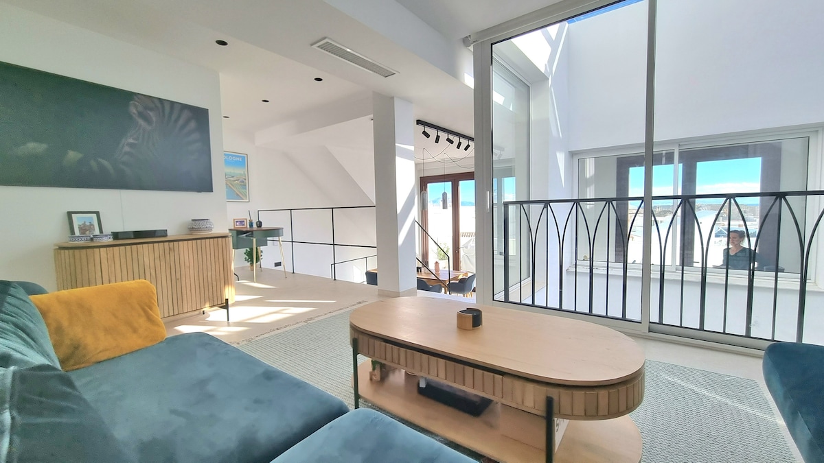 A comfortable living area features a spacious couch with colorful cushions, paired with a round coffee table. Large windows allow natural light to fill the room, enhancing the view of the outdoor terrace and distant landscapes. A subtle art piece is displayed on the left wall.