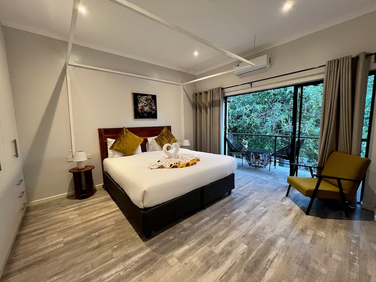 A spacious bedroom features a comfortable bed adorned with decorative pillows. Large glass doors open to a balcony surrounded by lush greenery, allowing for natural light. A seating area includes a chair next to the bed, enhancing relaxation in the room.