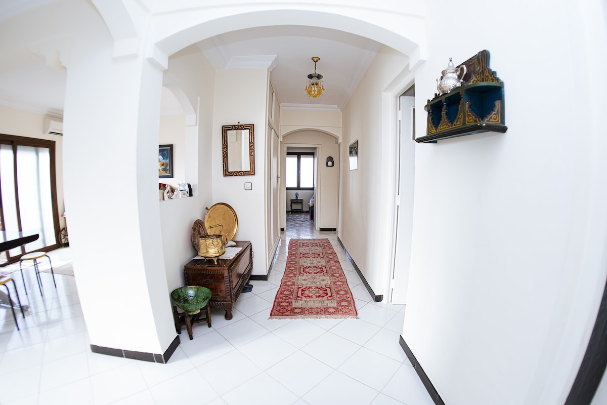 A bright hallway features tile flooring and white walls, creating an open atmosphere. Traditional Moroccan decor is visible, including a patterned runner and decorative wall pieces. Natural light pours in from windows at the far end, enhancing the welcoming feel of the space.