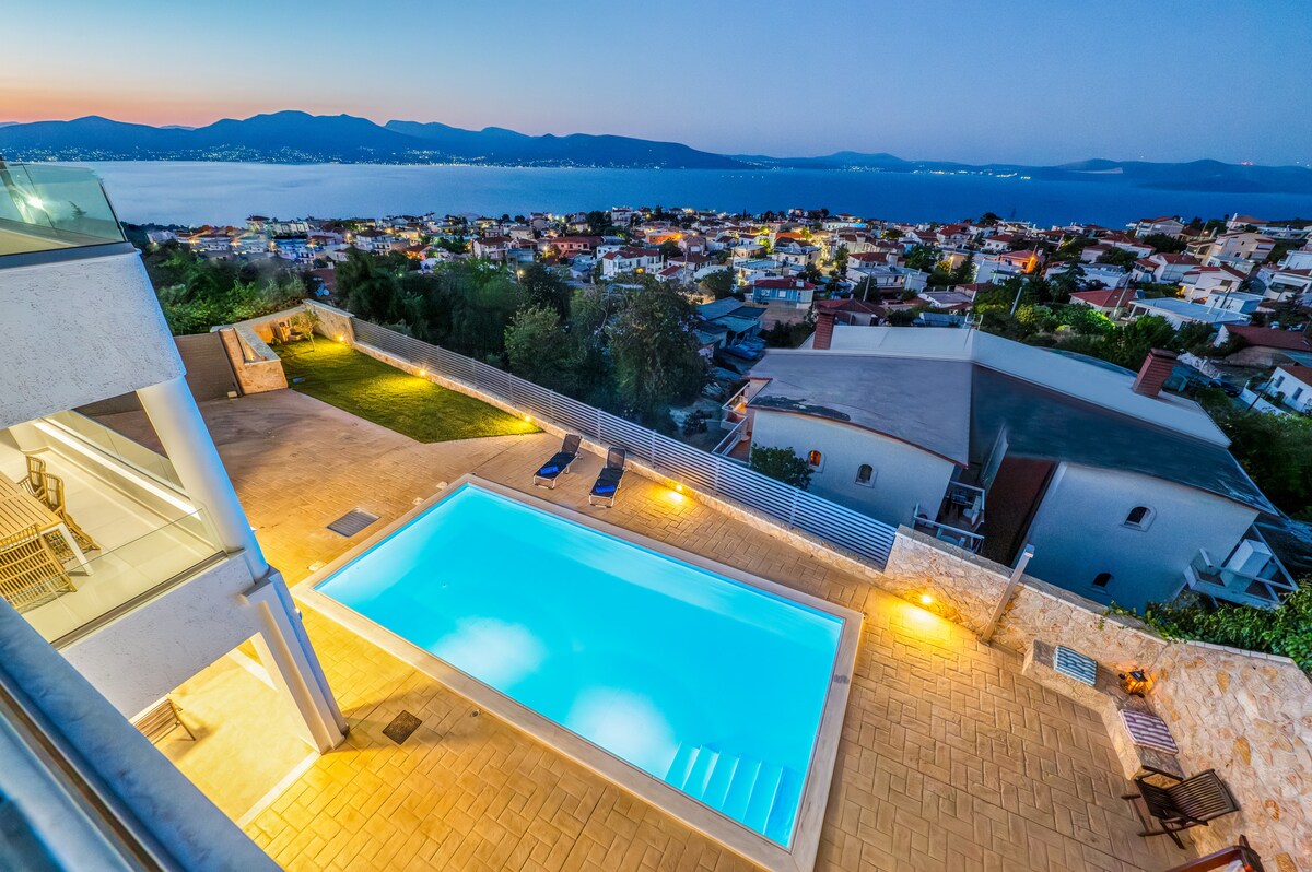 An outdoor area features a private pool illuminated by soft lighting, reflecting the surrounding landscape at dusk. Lounge chairs are positioned nearby, offering a view of the distant mountains and town below, creating a serene setting for relaxation.