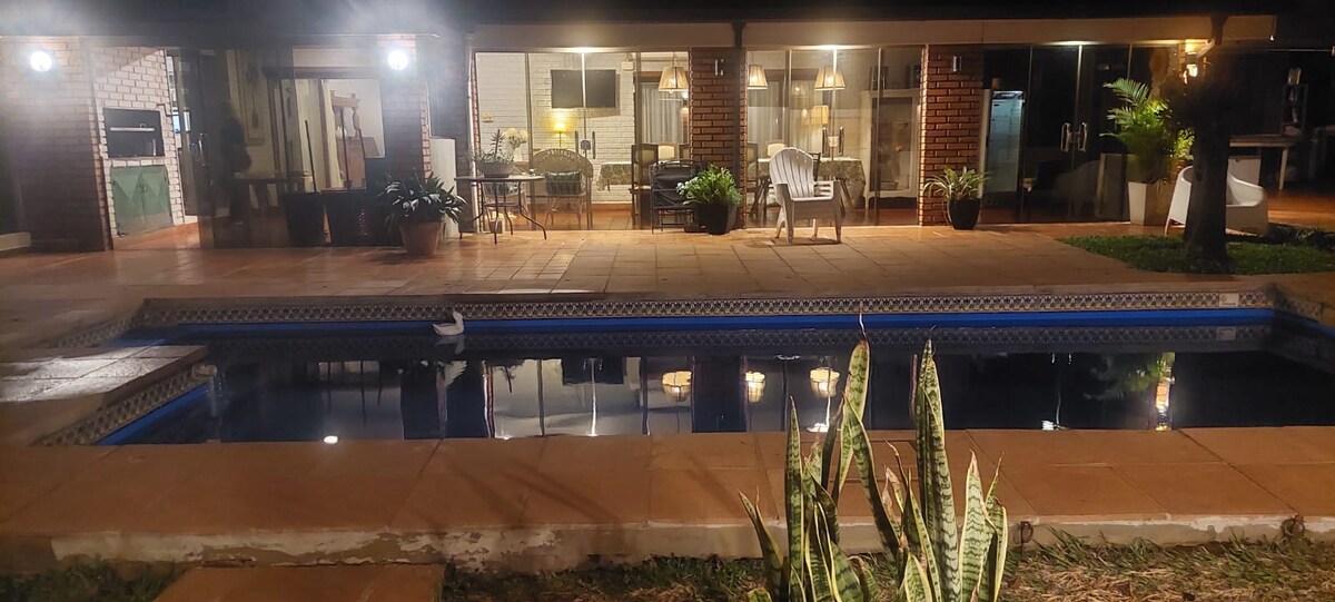 An outdoor area featuring a swimming pool surrounded by tiled decking. The pool reflects light from nearby lamps. In the background, a comfortable seating area and dining space are visible, highlighted by warm lighting. Lush greenery is present along the edges, enhancing the tranquil setting.