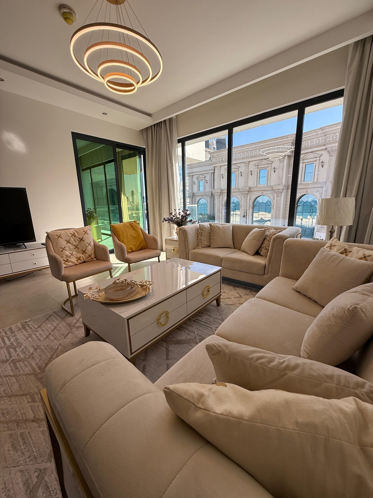 A bright living area features large floor-to-ceiling windows that allow natural light to fill the space. Stylish furniture includes a plush sectional sofa, two accent chairs, and a modern coffee table. Decorative elements and city views contribute to the elegant ambiance.