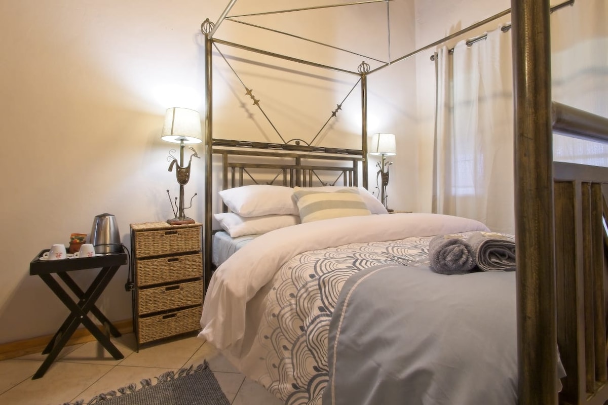 A cozy bedroom features a four-poster bed dressed in soft linens with a light blue duvet. A small bedside table holds a kettle and cups. Textured baskets offer additional storage. Gentle light filters in through soft curtains, highlighting the inviting space.