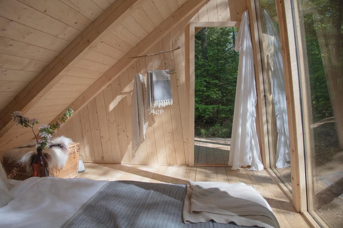 A cozy cabin interior features wooden walls and a sloped ceiling, allowing natural light to fill the space. A spacious bed is adorned with soft textiles. Nearby, a doorway with flowing sheer curtains leads to the forest, enhancing the connection to nature.