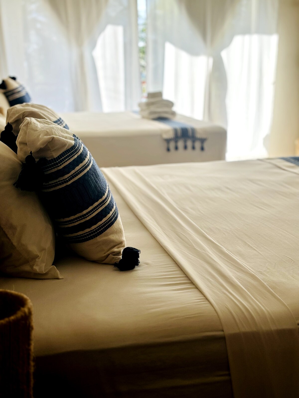 A calming sleeping area features two beds adorned with light-colored linens and decorative pillows with blue stripes. Soft, natural light enters through sheer curtains, creating a serene ambiance. A textured basket is positioned near the bedside, adding an element of comfort to the space.