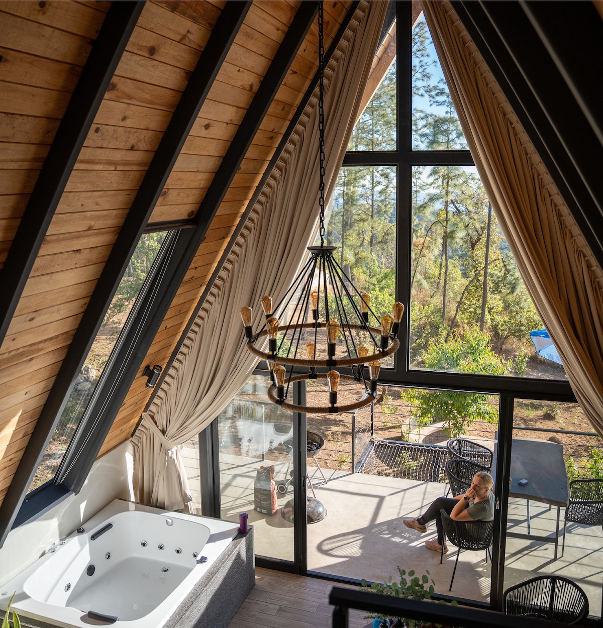 A tall A-frame structure features large triangular windows that fill the interior with natural light. A luxurious jacuzzi is situated in the corner, while a modern chandelier hangs from the vaulted ceiling. The outdoor area is visible through glass doors, offering views of the surrounding nature.