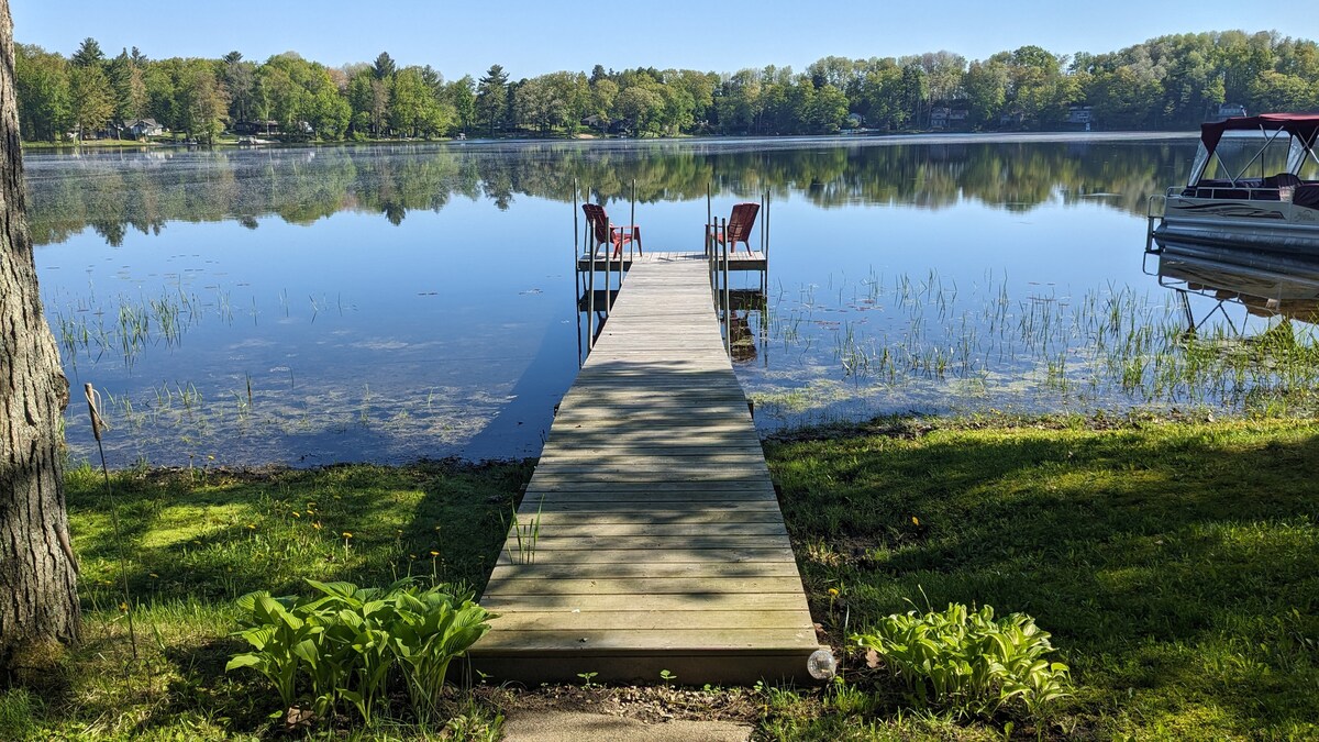 A wooden dock extends into a still lake, framed by lush greenery on both sides. Two red chairs are positioned at the end of the dock, offering a peaceful view of the calm water and surrounding trees.