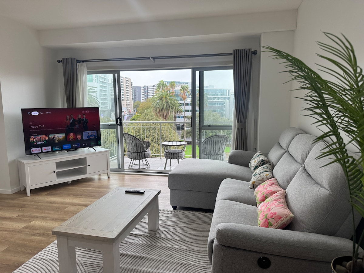 A modern living area features a spacious gray sofa equipped with decorative pillows, positioned adjacent to a stylish coffee table. A large smart TV displays streaming content, while sliding glass doors lead to a balcony with two outdoor chairs, providing views of the surrounding greenery.