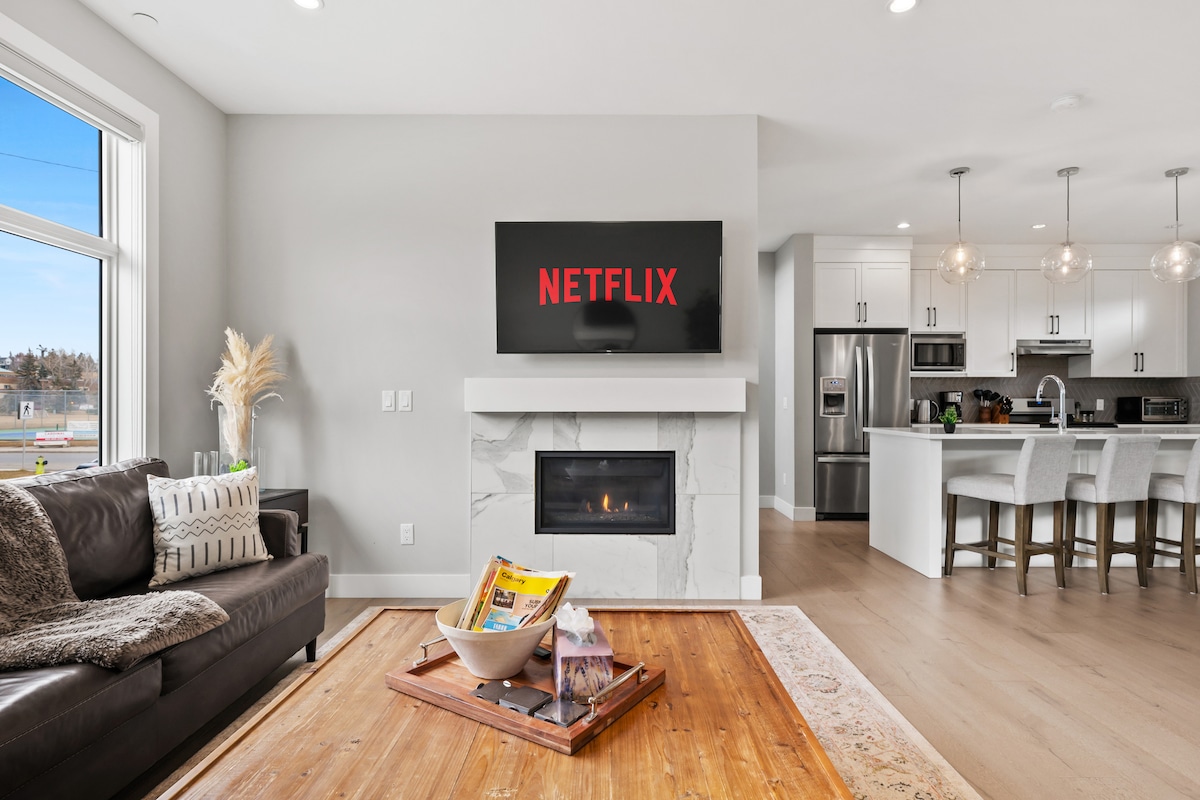 A modern living area features a comfortable leather couch set against a light wall. A large screen displays 'NETFLIX' above a stylish fireplace. A wooden coffee table holds magazines, while a bright open kitchen is visible in the background, showcasing sleek appliances.
