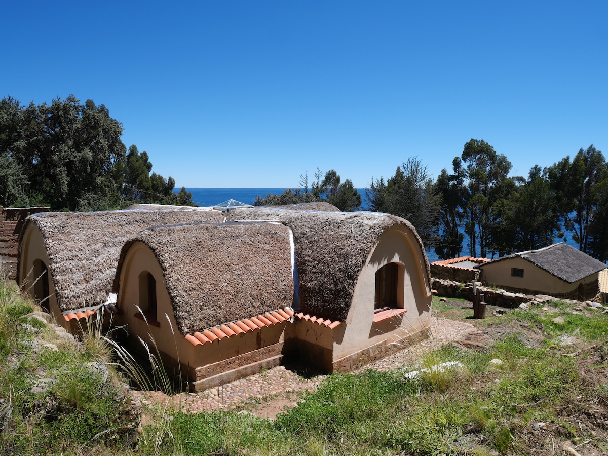A group of eco-friendly cabins is seen with thatched roofs, blending harmoniously into the natural surroundings. The cabins are positioned on a gently sloping landscape, overlooking a body of water. Clear blue skies and greenery frame the scene, creating a serene environment.