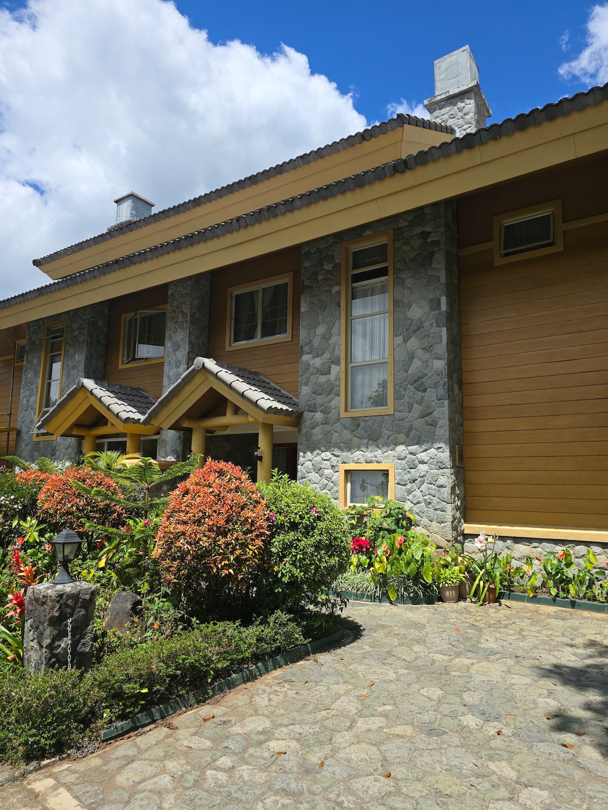 The cabin's exterior features a blend of wooden and stone elements, with a stone pathway leading to the entrance. Vibrant flowers and greenery surround the entrance, adding color to the setting. Large windows allow natural light to illuminate the interior.