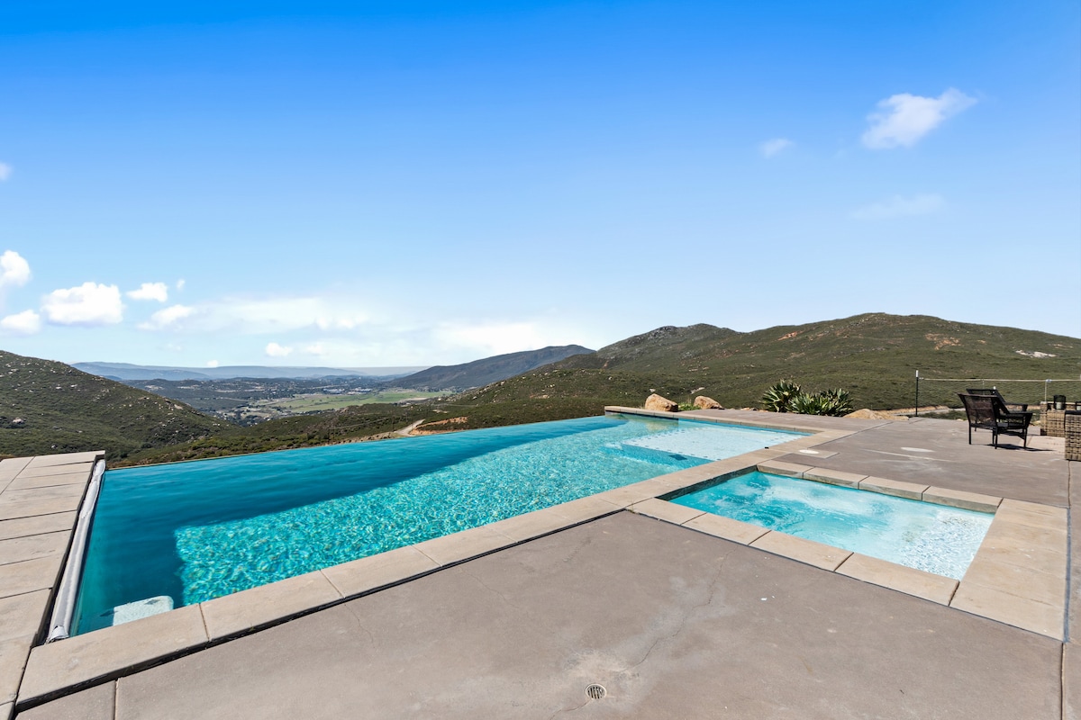 An infinity pool extends towards the horizon, offering stunning views of the mountains and valleys beyond. The pool area features a separate jacuzzi, both set on a spacious paved deck. Comfortable seating arrangements are visible nearby, enhancing outdoor relaxation.