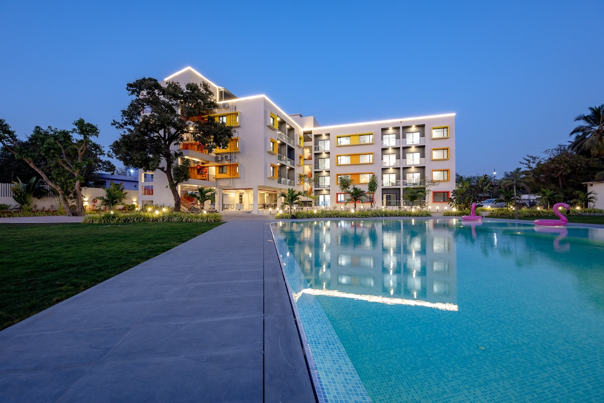 The exterior of the modern building is illuminated at dusk, displaying balconies along the facade. A tranquil swimming pool reflects the lights, surrounded by landscaped gardens and a spacious lawn. Palm trees and decorative lights enhance the welcoming atmosphere of the property.