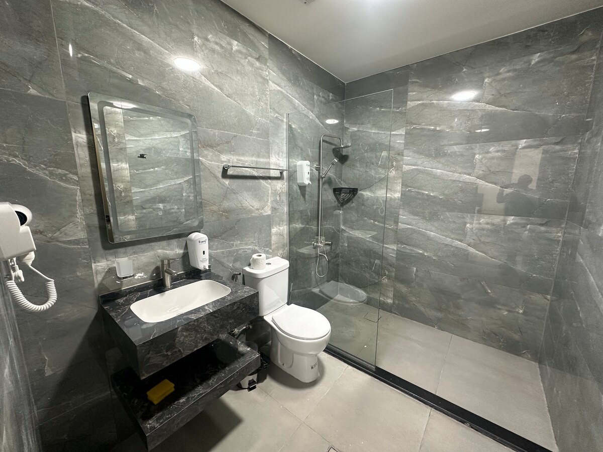 A modern bathroom is presented with sleek grey marble walls. A glass-enclosed shower is visible, alongside a white toilet and a dark countertop with a sink. A mirror hangs above the sink, with a hairdryer mounted nearby for convenience.