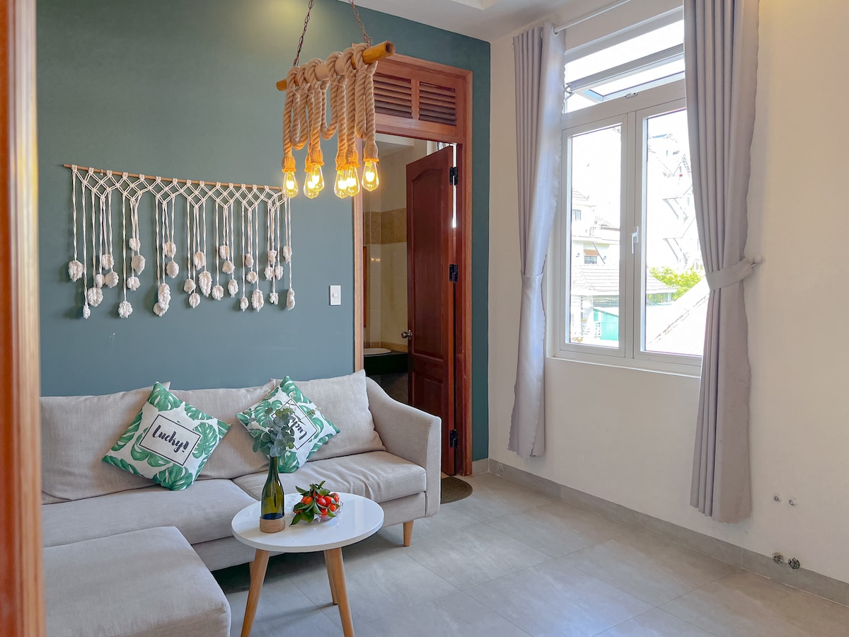 A bright seating area features a light-colored sofa adorned with green-patterned cushions. A hanging light fixture made of rope illuminates the space. A small table holds a flower vase and fruits, with natural light streaming through the nearby window.