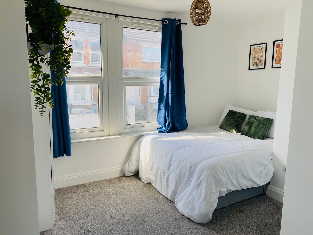 A bright sleeping area features a neatly made bed with white linens and green accent pillows. Natural light filters through two large windows with blue curtains, while a decorative plant and wall art enhance the serene environment.