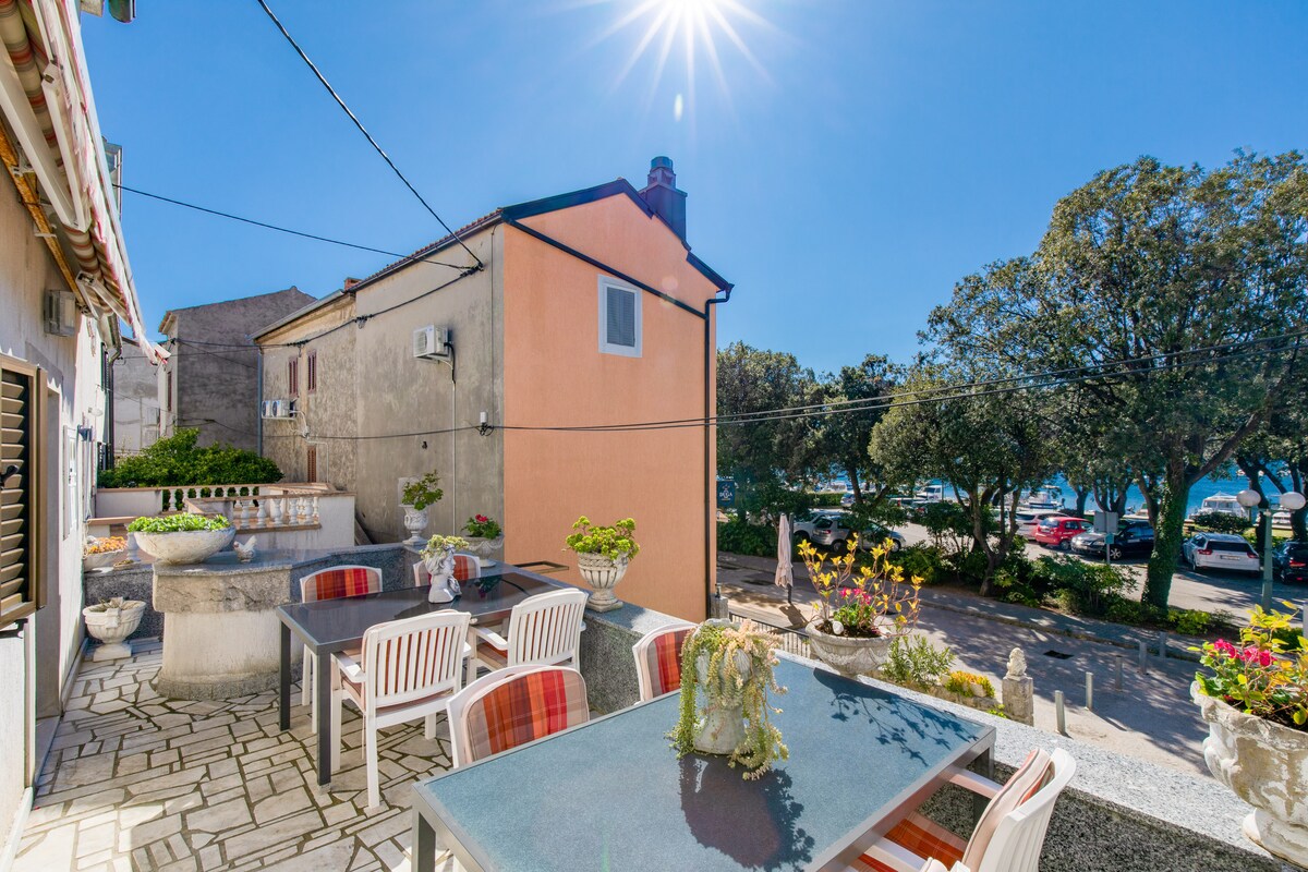 An outdoor terrace is shown, featuring a dining table surrounded by multiple chairs. Lush greenery and blooming flowers are visible, with the sea and promenade in the distance. The sun shines brightly overhead, enhancing the vibrant atmosphere of the space.