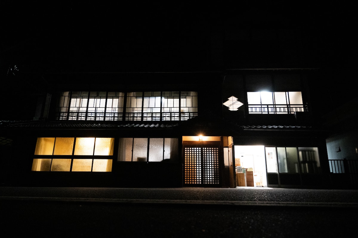 The traditional three-story wooden building is illuminated by soft light from its windows, creating a cozy ambiance. The entrance features a wooden lattice door, and the contrasting darkness highlights the structure's architectural details.