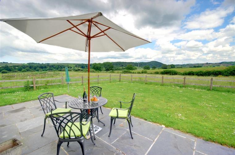 A shaded patio area features a round table surrounded by four chairs with vibrant green cushions. An umbrella provides protection from the sun, and the expansive lawn extends into scenic views of rolling hills under a partly cloudy sky.