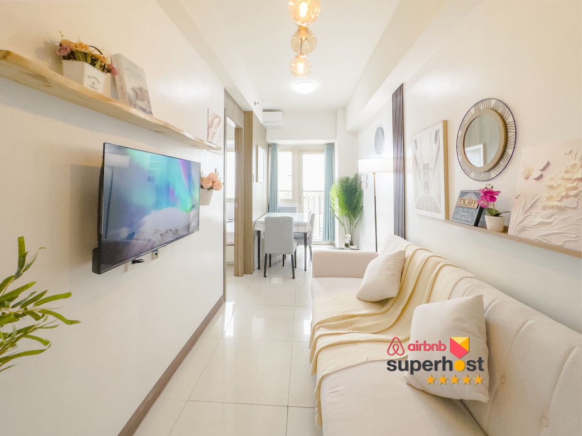 A cozy living area features a light-toned sofa and a mounted television displaying calming visuals. Natural light filters through the window, illuminating a small dining space in the background. Decorative elements, including framed art and a plant, add warmth to the environment.