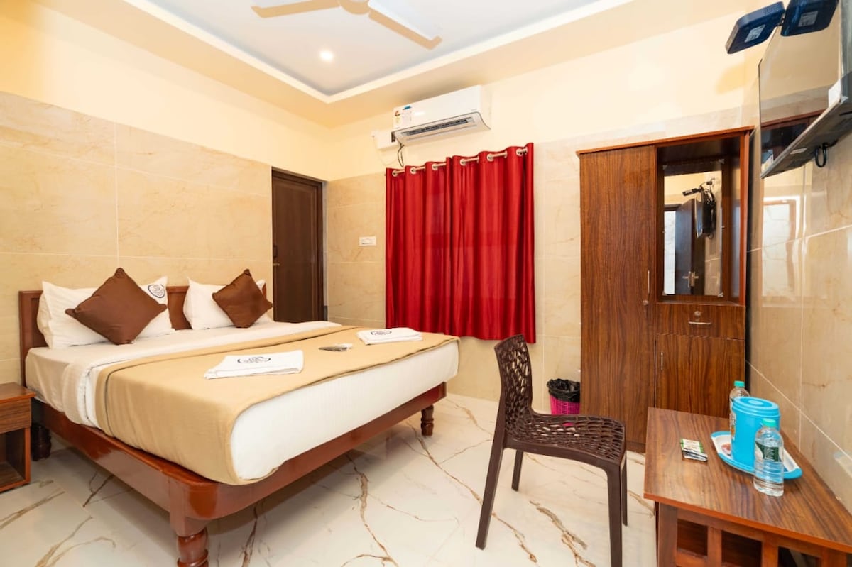 A well-lit room contains a spacious bed adorned with neutral bedding and two brown accent pillows. A wooden chair and table are present, with a water bottle and glass resting on the table. Red curtains frame the window, and a mirrored wardrobe is visible.