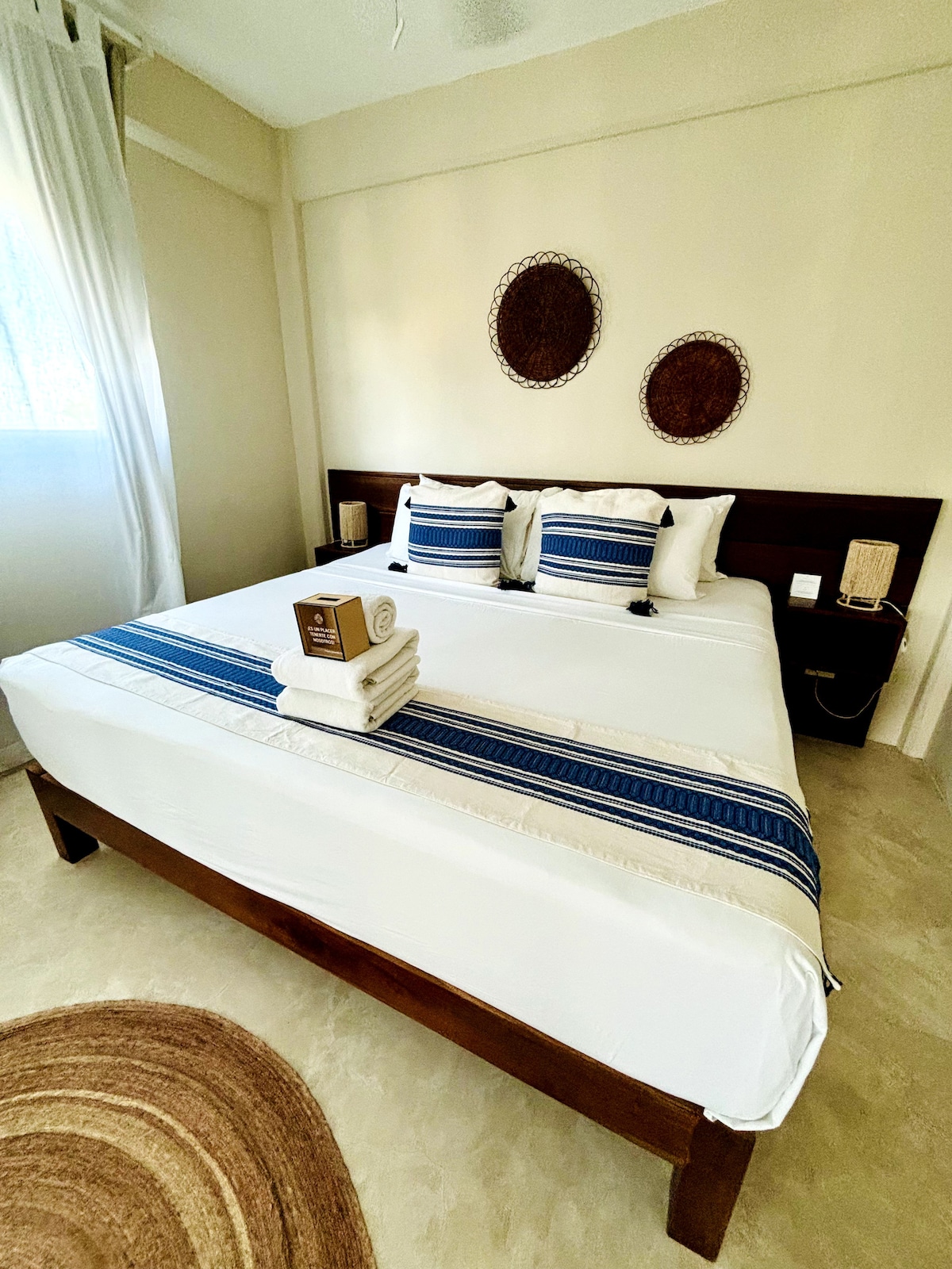 A spacious sleeping area features a large bed adorned with crisp white linens and a decorative blue and beige bed runner. Two circular woven wall hangings complement the neutral walls. Natural light filters through sheer curtains, illuminating the room's serene ambiance.