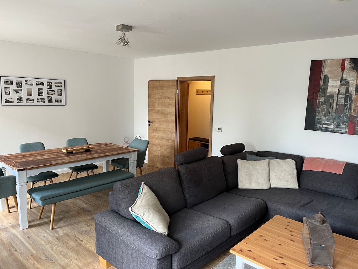 A living area is shown featuring a spacious sectional couch with accent pillows and a wooden coffee table. A dining area with a long table and six green chairs is visible in the background. The room has neutral walls and a piece of artwork on one side.