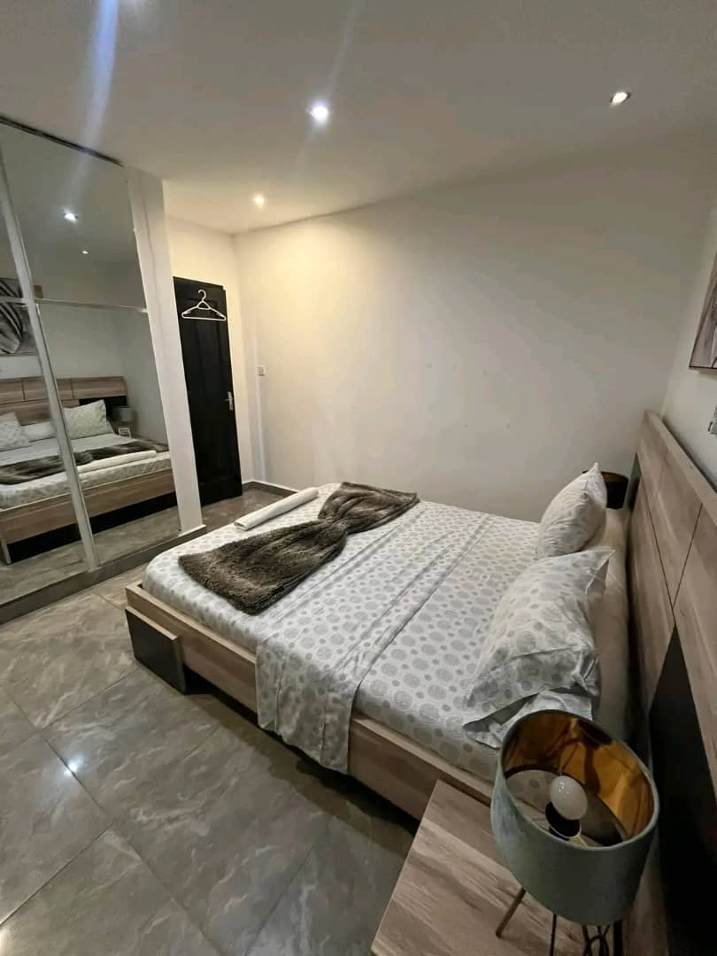 A one-room apartment features a spacious bed with a light-colored patterned duvet. A soft blanket is draped casually across the bed. A mirrored wardrobe reflects the room's neutral tones, while a bedside table with a lamp adds a touch of warmth.
