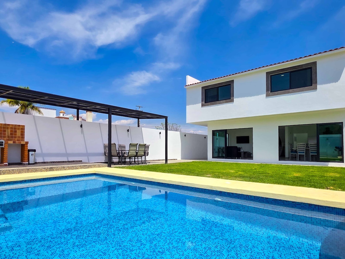 A modern two-story house is depicted, accompanied by a sparkling pool. The outdoor area includes a shaded dining set and a grassy lawn, providing ample space for relaxation. Large glass doors open into the living space, inviting natural light into the interior.