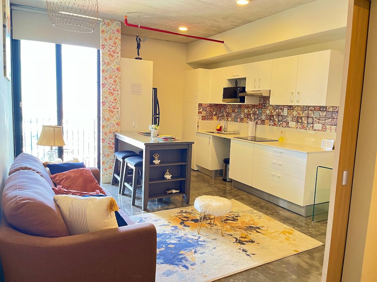 The living space features a cozy sofa adorned with cushions, a small white ottoman, and a colorful area rug. An open kitchen with modern appliances is visible, complemented by a dining table. Large windows allow natural light to fill the space.