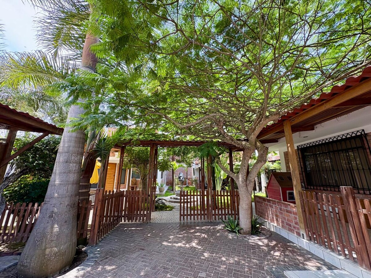 A welcoming entrance is framed by a lush green canopy provided by a large tree. A wooden fence surrounds the area, leading to a pathway that invites exploration. The surrounding structures hint at an engaging outdoor space, complemented by well-maintained greenery.