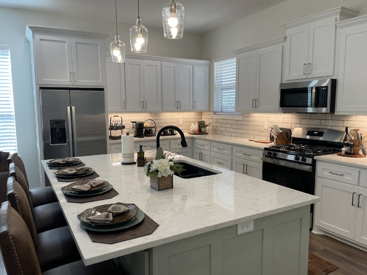 A modern kitchen features light cabinetry and stainless steel appliances. An island with seating for four is accompanied by elegant place settings. Pendant lights hang above, illuminating the space. Natural light enters through two windows adorned with blinds.