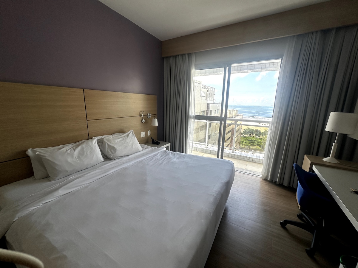 A bright sleeping area features a spacious bed with crisp white linens. Natural light is welcomed through a large glass door that leads to a balcony, providing views of the ocean. A desk and chair are positioned near the window, offering a functional workspace.