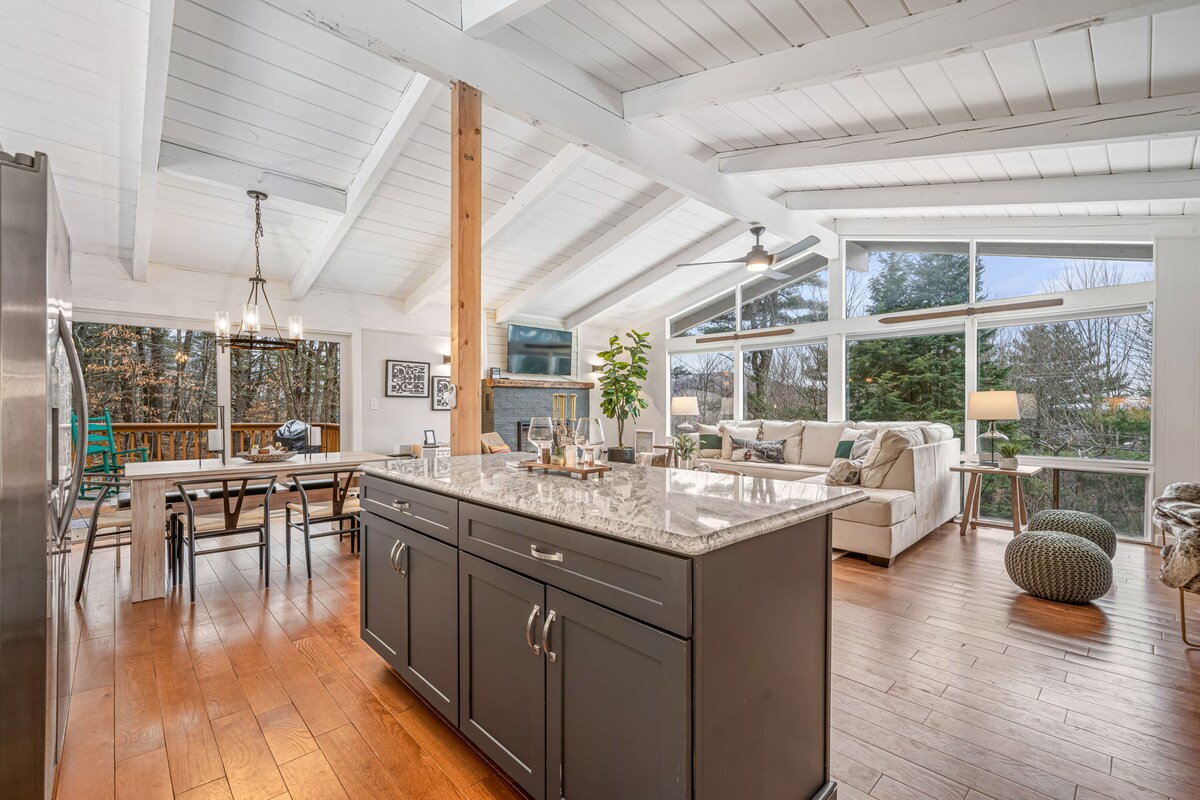 An open-concept living space features a modern kitchen with dark cabinetry and a granite island. A dining table and comfortable seating area with large windows create a bright atmosphere. Natural light illuminates the wood flooring, while decorative elements enhance the contemporary design.